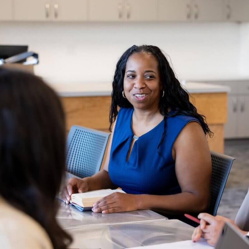 A group of educators sitting around a table in a school meeting space, smiling and engaged in collaborative conversation during a staff planning or leadership discussion.