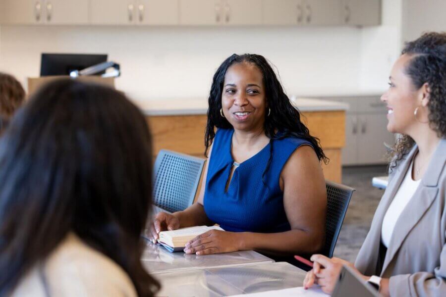 A group of educators sitting around a table in a school meeting space, smiling and engaged in collaborative conversation during a staff planning or leadership discussion.